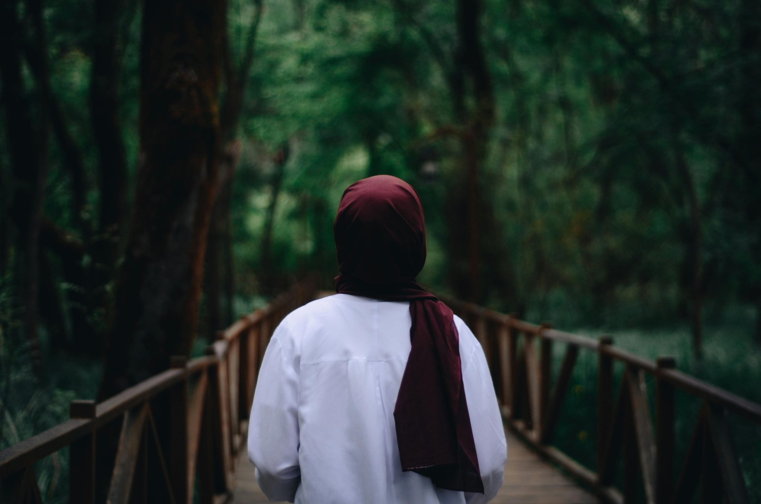 A woman in a hijab walks on a wooden bridge surrounded by lush greenery in a tranquil forest setting.